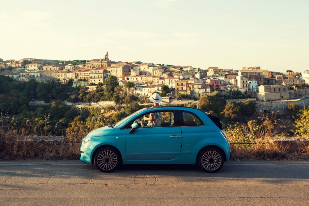 Woman enjoying a drive in a cute blue car overlooking a gorgeous Sicilian town at sunset