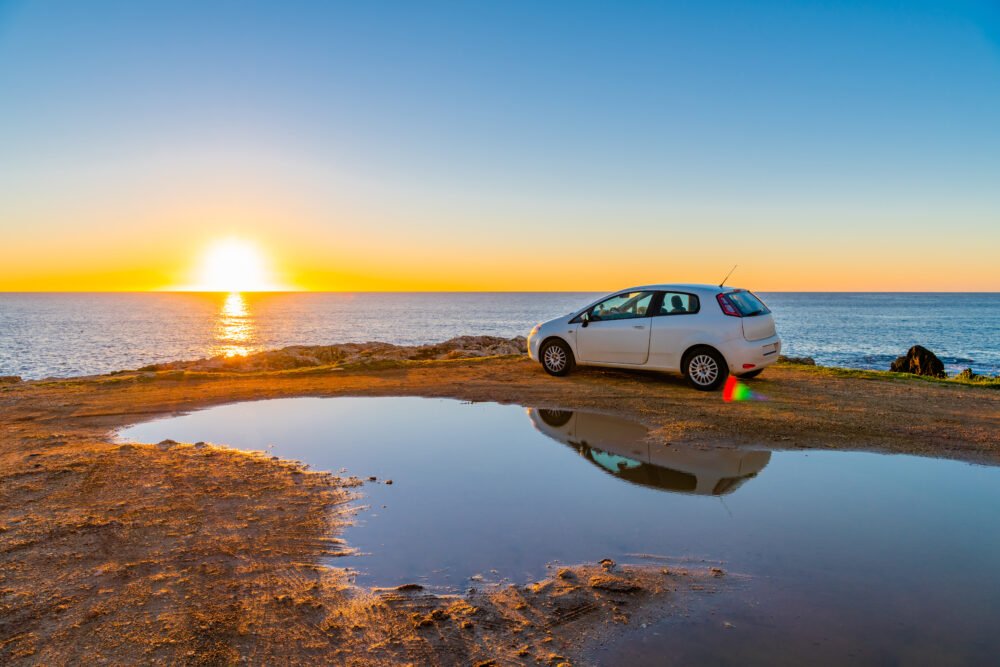 A stunning remote beach in Sicily at sunrise, with a white rental car at the edge of the road, as the sun sinks into the horizon with no one else around.