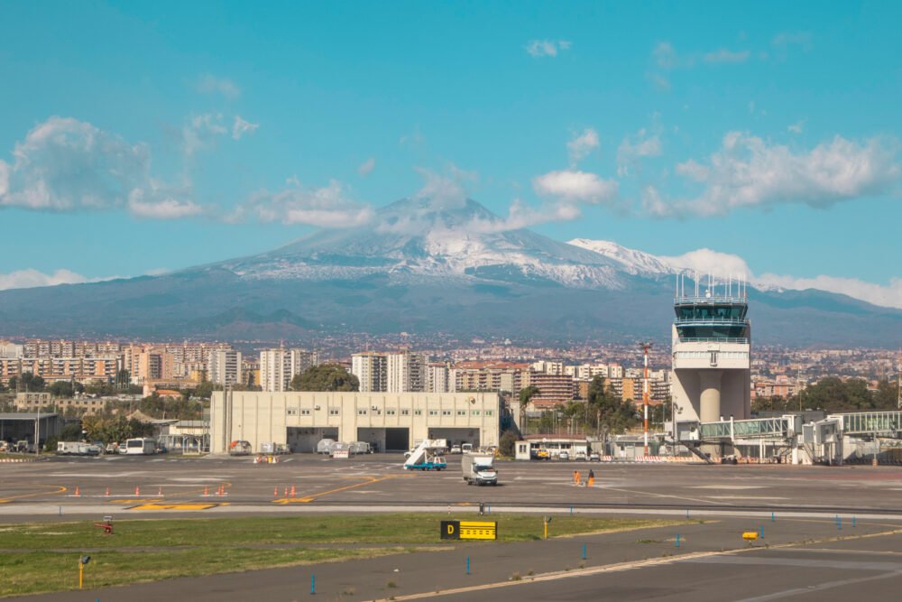 Catania Airport, in Sicily, seen with Mount Etna in the far distance, behind, on a clear day with just a few clouds in the sky. You can see the runway, a few cars and trucks, people, and air traffic controller, and then the city center in the background.