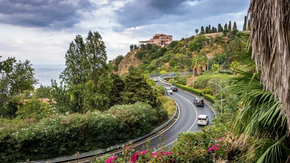 Road surrounded by green trees and plants with cars driving in Taormina, Sicily, with just a few other cars on the road on a cloudy day