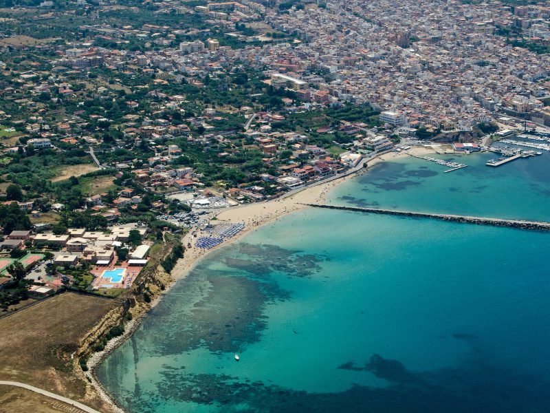 Aerial view of a beach near Palermo, Italy, which shows a beautiful turquoise shallow wading area and darker waters, as well as a pier stretching out from the ocean. The photo is taken from an aerial angle, perhaps from a drone or from a plane, and you can see the city below it.