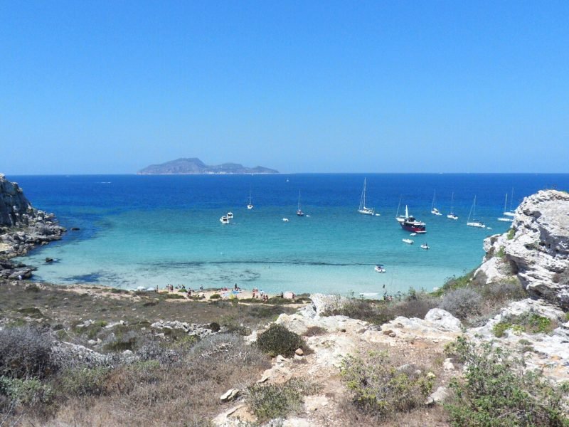 Small but beautiful strip of sandy beach leading out into crystal clear water with boats in the shallow water, with an island visible in the distance