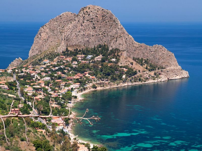 The rocky backdrop of Capo Zafferano with brilliant blue sea behind it and a strip of beach and houses on the cliffsides