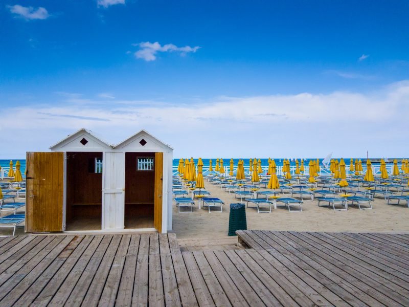 Two small changing rooms, with blue beach chairs and yellow umbrellas ready for the beachgoers who will be visiting later that day, and brilliant blue water in the background