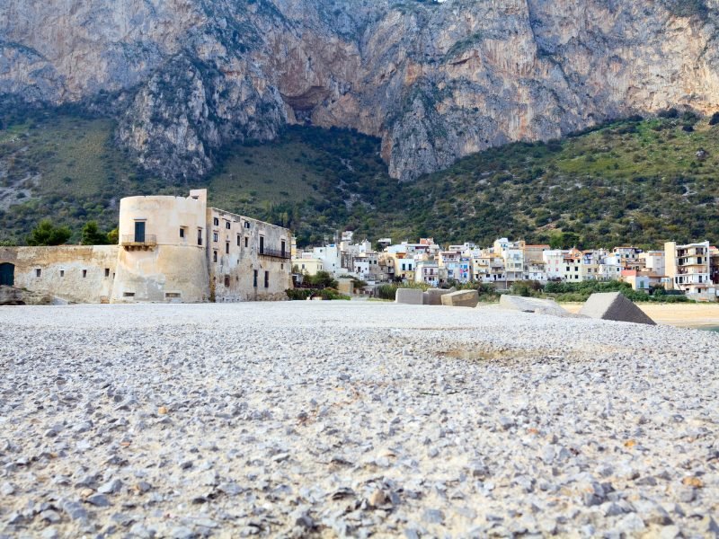 White, rocky sandy beach of Spiaggia Vergine Maria, with castle remnants and houses against a mountain backdrop