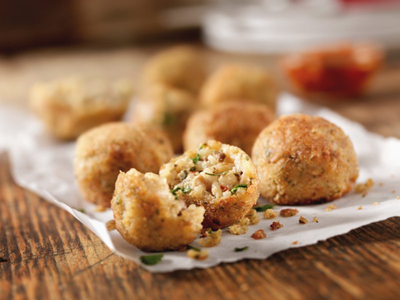 A selection of tasty arancini, fried balls of risotto stuffed with filings and herbs, one broken open to show interior, served on a wooden table with wax paper beneath it.