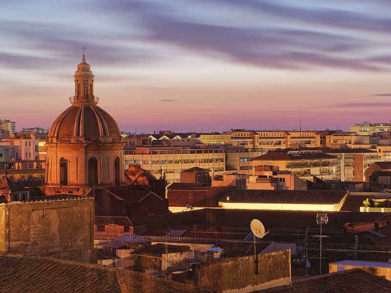 Sunset over the city of Catania, with pink and orange colors in the horizon, dome of a church, buildings of the city center visible