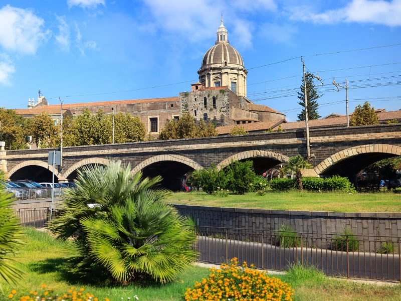 View of a bridge and parking area in Catania, with the church and an old city wall visible from the road