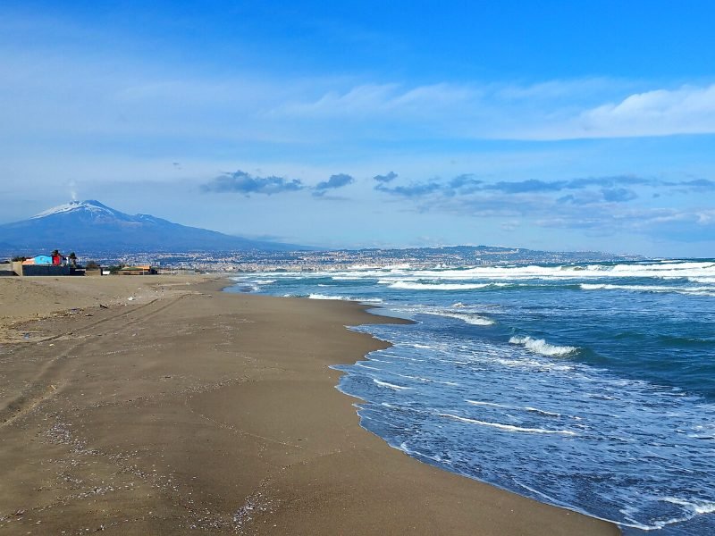 View of the Playa Catania with the city in the background and Mt Etna in the distance with snow and a small amount of smoke coming out of the top of the crater