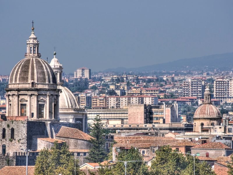 View of the churches and many large buildings of Catania, with the background of Mt Etna volcano in the distance, sloping upwards.