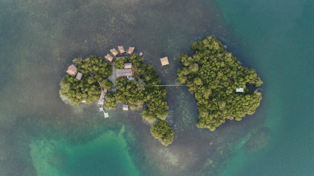 A drone shot showcasing the landscape of this private island, which consists of a handful of private overwater bungalows and a bridge to another small island that has very minimal development. The water around the islands is dark colored where there is some reef, and then deep turquoise further out.