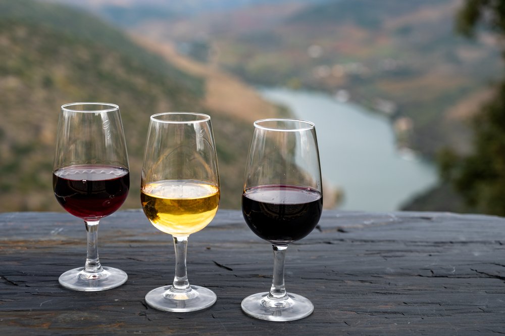 Three glasses of wine, red, white, and sweet port, on a wooden table overlooking the Douro River behind it