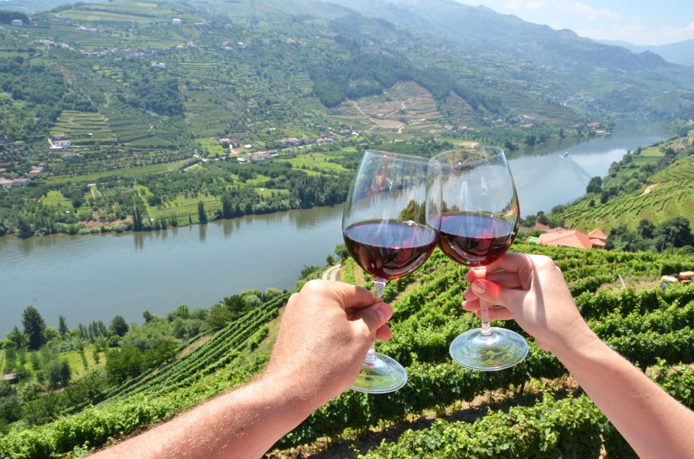 Two people clinking their wine glasses together, full of red wine, with a view of the Douro River and vineyards of the Douro Valley in the background