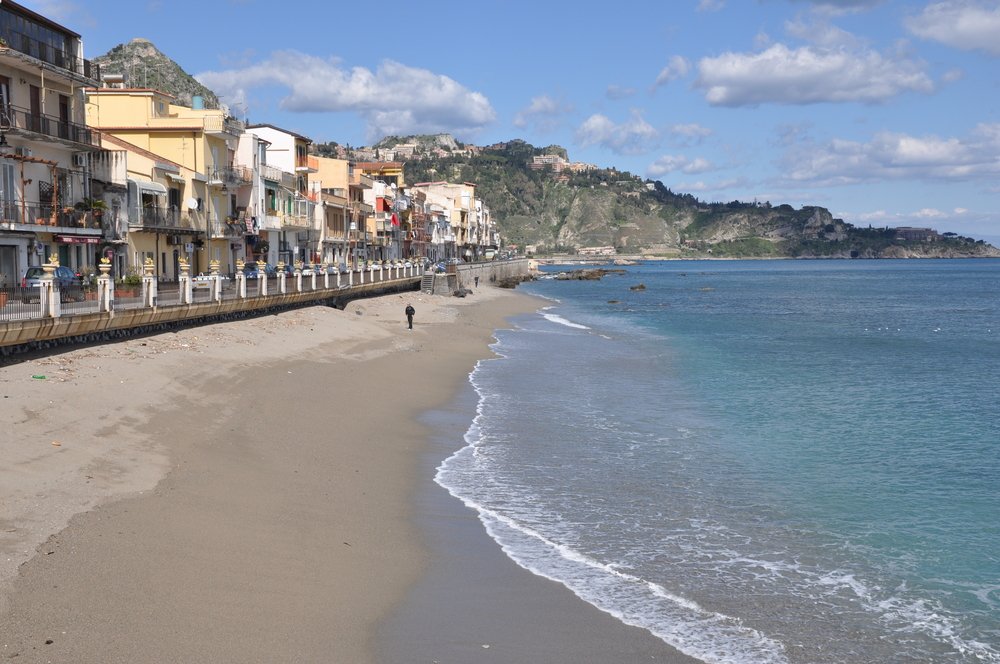 Beach in Giardini Naxos, Sicily with beachfront, one person on the beach, hillside mountains and clear waters