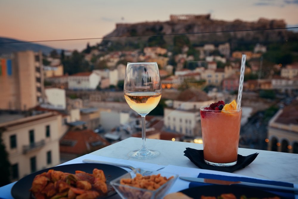 Greek rooftop view with wine, cocktail, greek food and a blurry view of the acropolis on the hill in the background