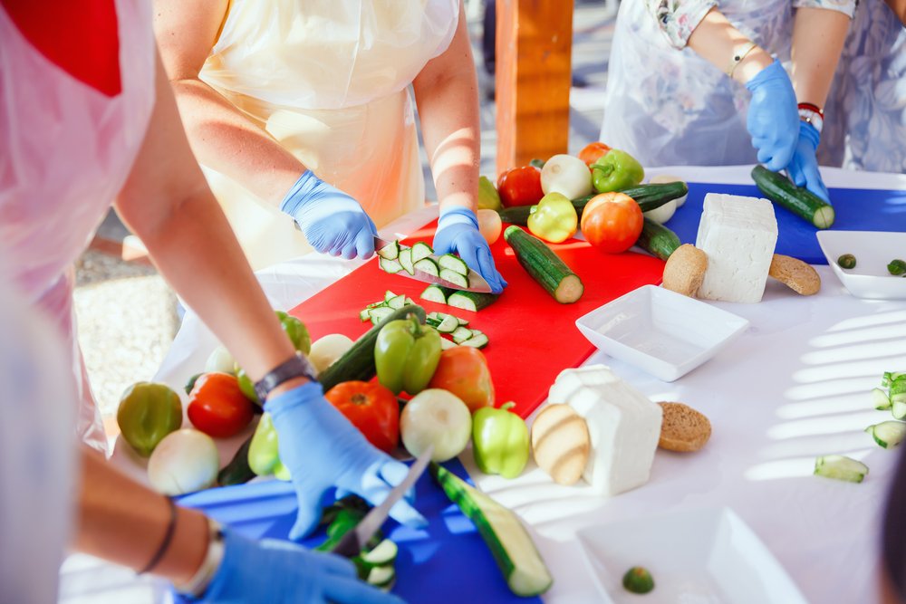 Several womans' hands cutting vegetables like tomatoes, bell pepper, onion, etc. for Greek salad at a cooking class in Athens