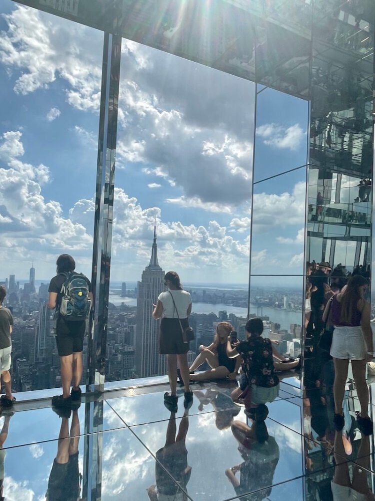 the mirror room in the popular summit one observation deck in nyc with gorgeous views of the empire state building on a sunny summer day with clouds and skyline reflecting on the mirrored floor