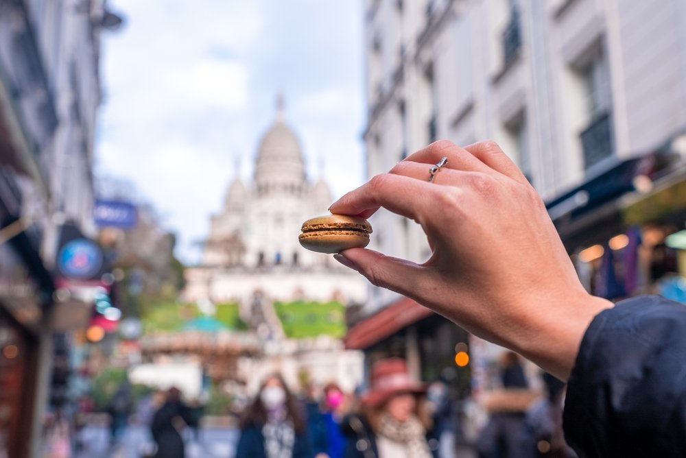 Person's hand holding a macaron cookie in front of a Parisian landscape