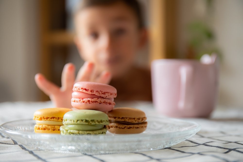 Child reaching for a plate of macarons, in focus, pink yellow green and brown cookies, and a pink mug visible too.
