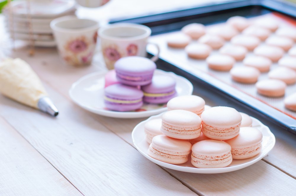 Colorful pastel pink and purple macarons with two floral tea cups, pastry bag, and cooked macaron shells on a baking sheet