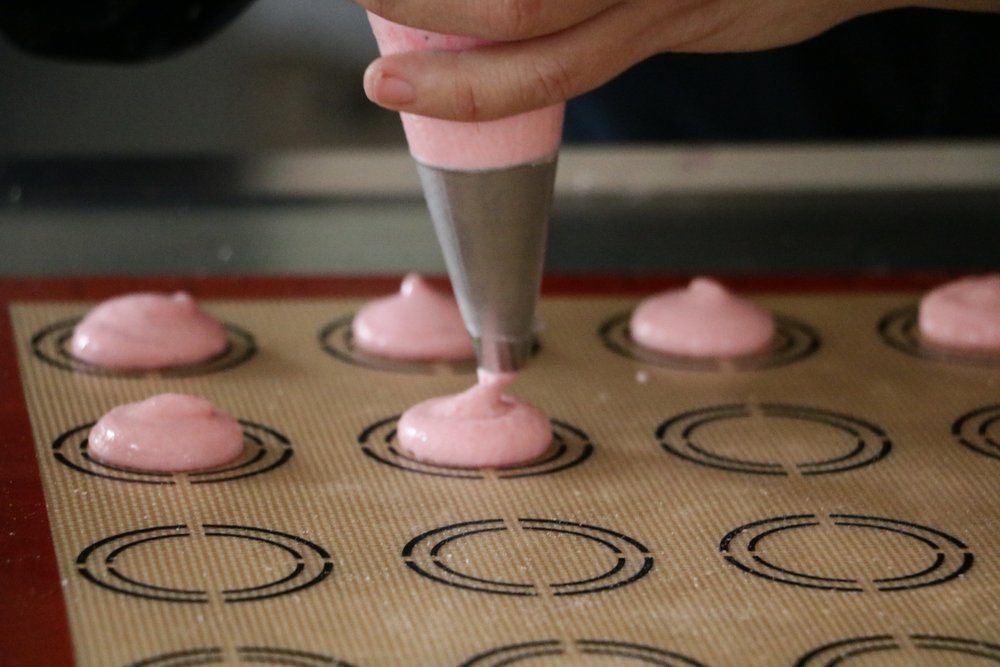 Person using a pastry bag to pipe macarons onto a baking sheet with circles on it to indicate the size that the macaron batter should be