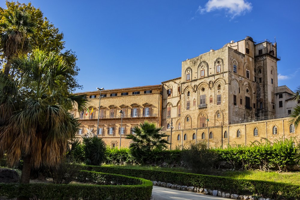 Norman Palace - one of oldest royal palaces in Europe; it was created in IX century by Emir of Palermo, with palm trees and foliage in front of a beige castle-like palace