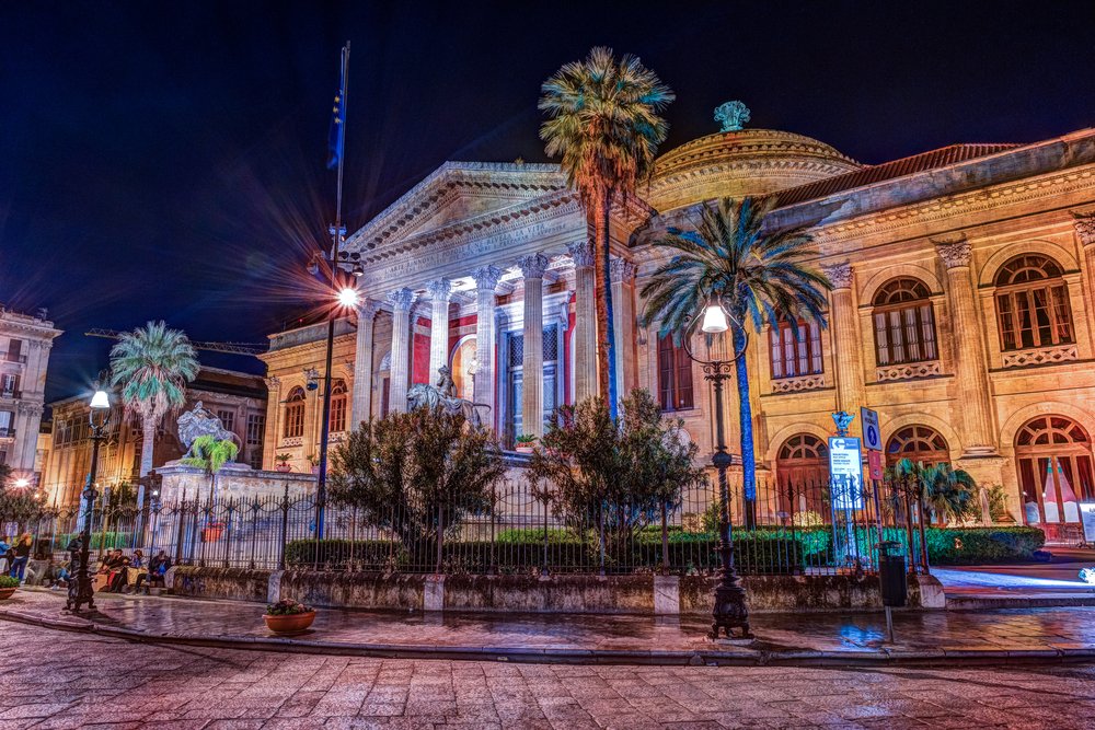 The evening view of Teatro Massimo - Opera and Ballet Theater in Verdi Square, Palermo, Sicily, with lights and palm trees in front of it