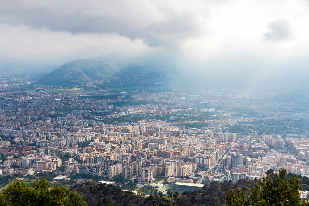 View of Palermo as you start to fly into its airport, half an hour from the city center, to start your Palermo itinerary. Hazy clouds, city buildings, mountains in the background.