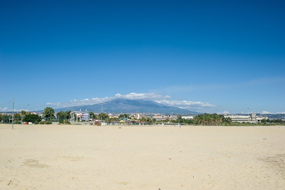 Soft pale sandy beach with Mt. Etna in the background and clouds on the top of the volcano on a sunny day on a Catania beach
