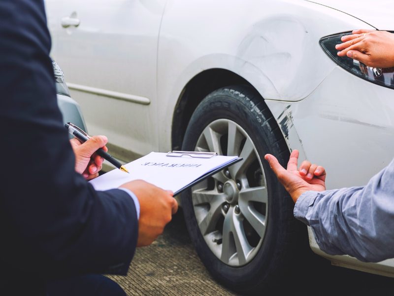 Two men discussing car insurance after an accident where a white car shows mild damage to the front of the car.