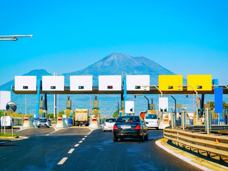 Cars making their way to different lanes at a toll booth area before the autostrada in Catania, Italy, with Mt. Etna visible in the background on a clear day.