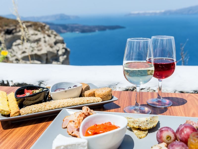 two glasses of wine at a winery in santorini with a beautiful view and a small plate of things to nibble on while you drink wine