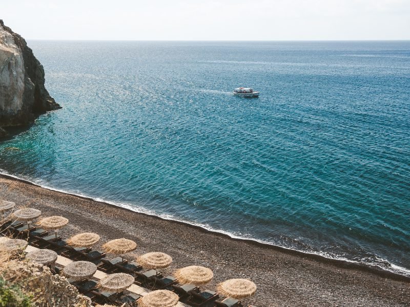 a beautiful black sand beach in santorini with umbrellas unused because it is early in the day and a boat out on the santorini mediterranean water