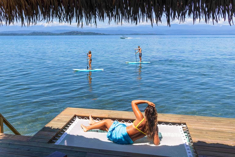 Woman sitting on net of a deck watching two paddle boarders in Panama