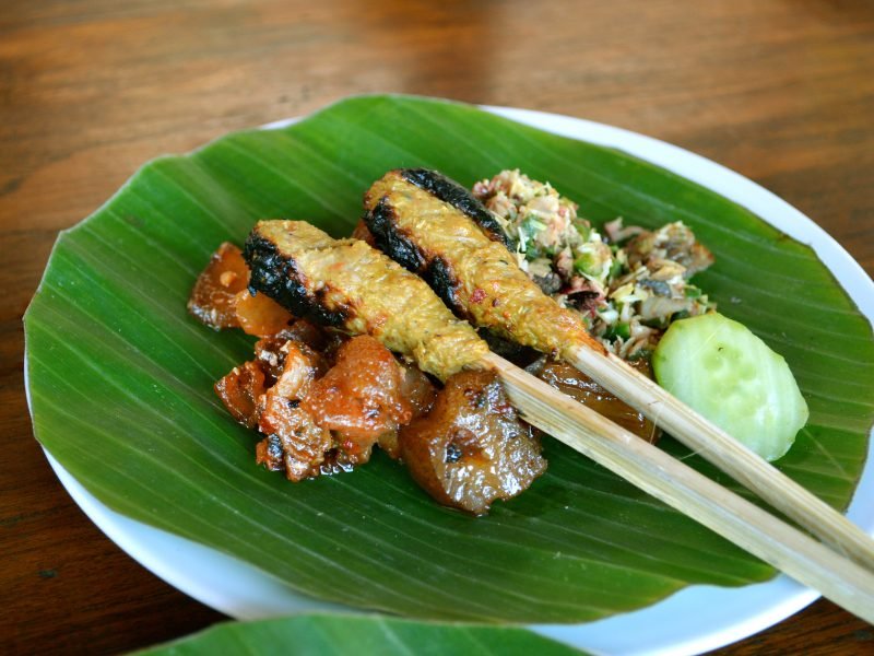 Minced chicken satay on bamboo skewers served on a leaf plate with cucumber and samba