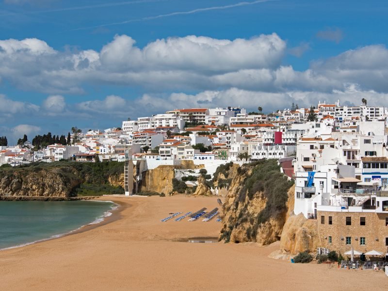 the beach on the town of albufeira with whitewashed houses, dark yellow sand beach, cliffside hosues, and clear waters