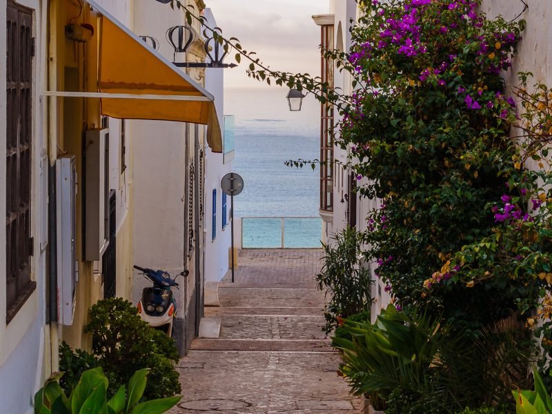 view of albufeira old town with scooter, steps, and bougainvillea, and sea in the distant background.