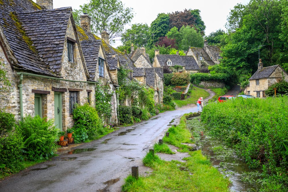 a road in the cotswalds with stone houses and bushes and small street