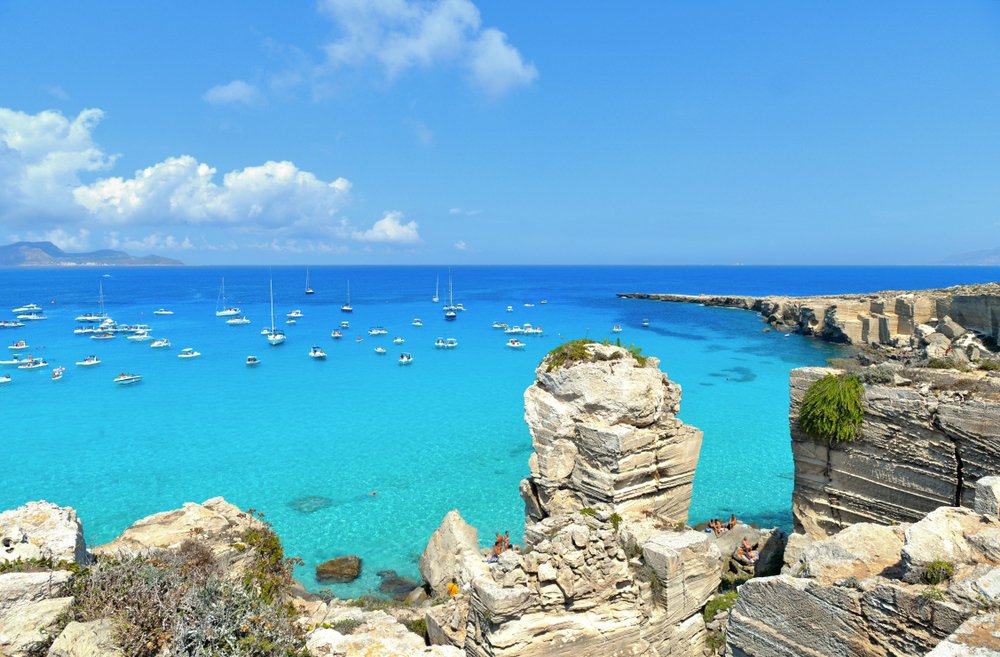 The beautiful turquoise waters of Cala Rossa beach with an island in the background, lots of boats in the water