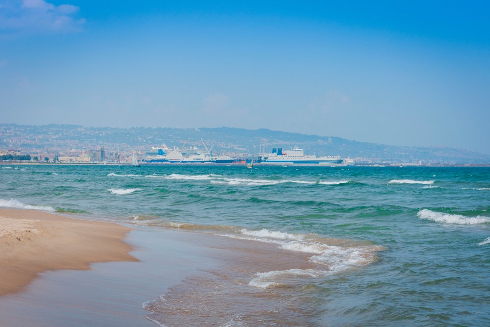 The Catania-area beach of Lido Azzurro with ships, waves, and city in the background, with soft beige sand that is pleasant to walk on