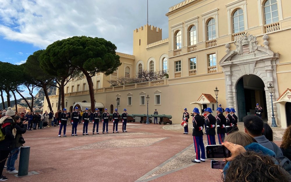 Tourists gathering and holding out their cellphones to record the daily changing of the guards ceremony which takes place in front of the pastel yellow Prince's Palace of Monaco. There are about 12 guards in two rows of six facing each other. The weather is sunny.