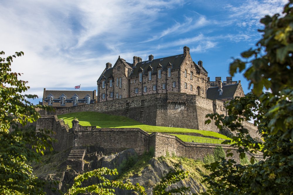 View of the Edinburgh Castle from up on a hill, looking up from below, on a sunny day with green grass surrounding the castle.