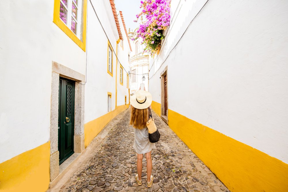 Blondish-brown haired woman with a white hat in the white and yellow streets of Evora, Portugal with pink flowers blooming and cobblestone streets