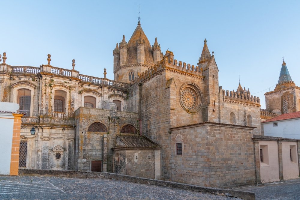 Morning light on this view of the cathedral in Portuguese town of Evora. The cathedral is made of tan stones with a circular stained glass window and lots of castle-like design elements. There is no one there and the sky is clear.
