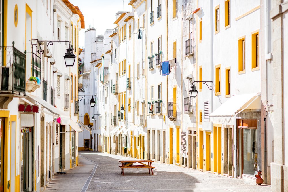 Street view of the residential buildings in Evora City with white walls and colorful yellow windows and door frames and a bench in the middle of the street