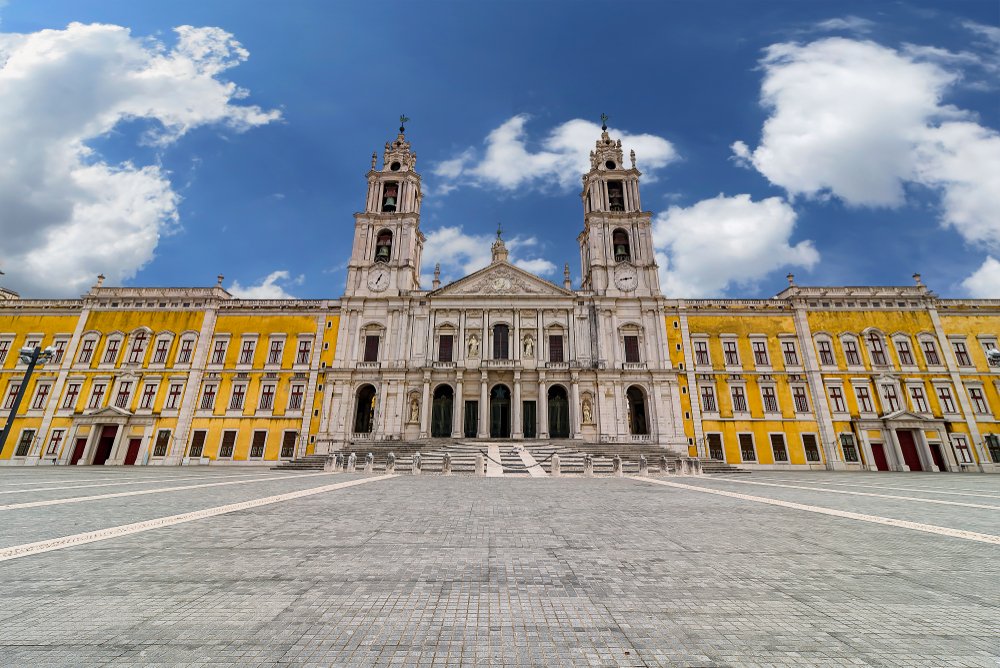 View of the yellow and gray palace, with an ornate facade and two towers with clocks on them, on a sunny day with no one around in front of the palace.