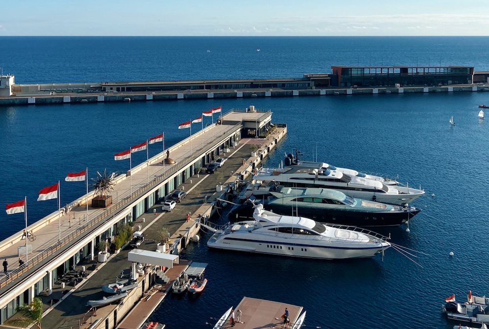 The Monaco marina of Hercules Port. You can see the Monaco flag which is red and white showing, and a handful of beautiful speedboats docked at the marina. The sea is azure and calm and it is a beautiful day.