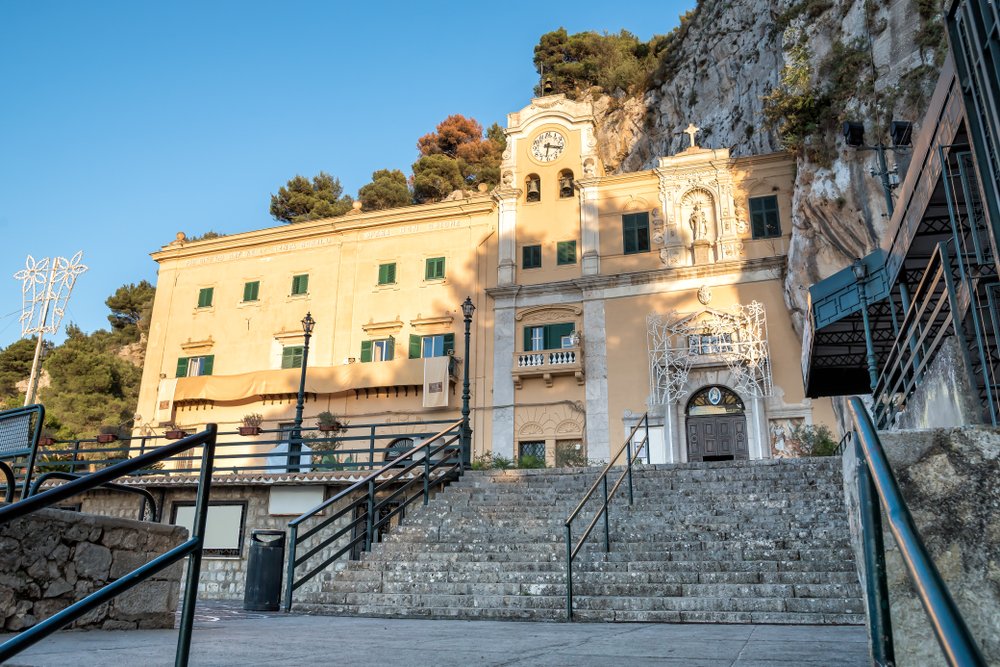 View of Sanctuary of Saint Rosalia with the holy cave on top of Monte Pellegrino in Palermo, Sicily, a yellow-toned church with a clock built into the side of a mountain near Palermo.