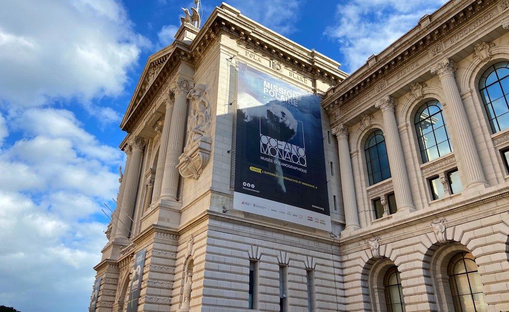 The striking white building of the Oceanographic Museum of Monaco, which hosts an aquarium. The building has many pillars and arched windows and looks like a palace. There are some clouds in the sky but it is mostly sunny.