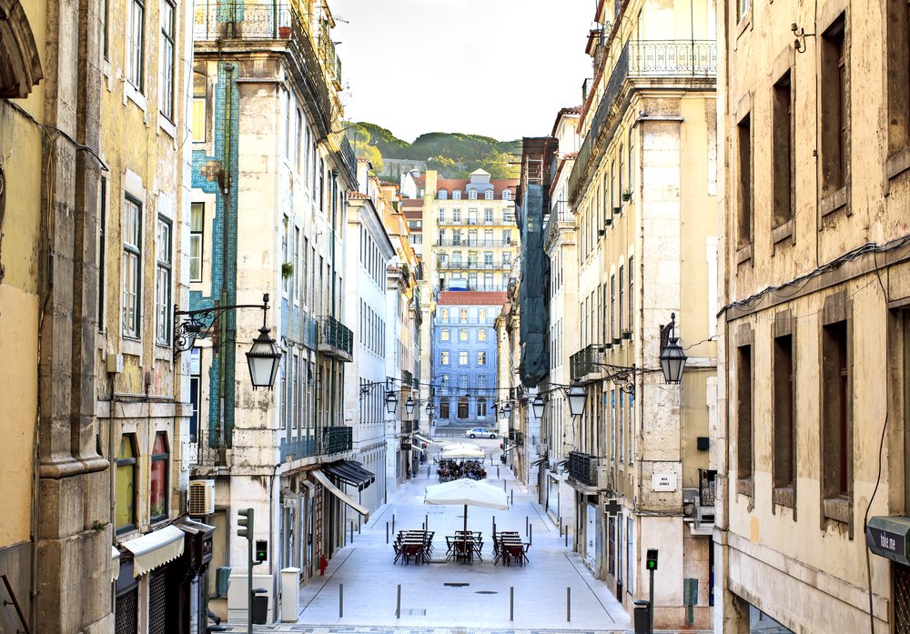 Vew from the exit of the metro station "Baixa" with some open-air seating in the street, and views of buildings stacked on a hillside in the distance.